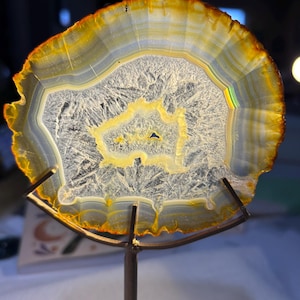 May include: A translucent, circular agate slice featuring concentric bands of yellow, amber, and white, mounted on a simple metal stand. The center displays a crystalline, textured cavity with a small, dark, irregular opening in the middle.