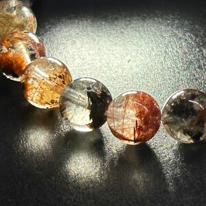 Puede incluir: Primer plano de una pulsera con cuentas redondas y transparentes. Cada cuenta tiene inclusiones doradas y rojizas. Las cuentas est&aacute;n ensartadas, creando un efecto brillante. Fondo oscuro y reflectante.