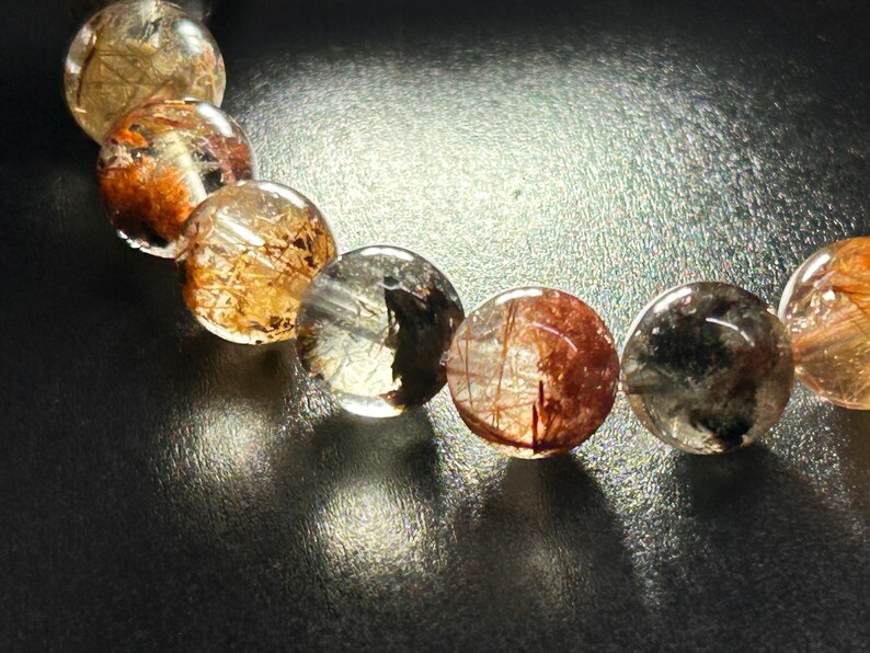 Puede incluir: Primer plano de una pulsera con cuentas redondas y transparentes. Cada cuenta tiene inclusiones doradas y rojizas. Las cuentas est&aacute;n ensartadas, creando un efecto brillante. Fondo oscuro y reflectante.