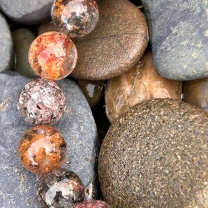Puede incluir: Primer plano de varias piedras preciosas esf&eacute;ricas transparentes con inclusiones internas de color naranja, marr&oacute;n y negro. Las piedras preciosas est&aacute;n dispuestas entre cantos rodados lisos, grises y marrones, creando una est&eacute;tica natural y terrosa.