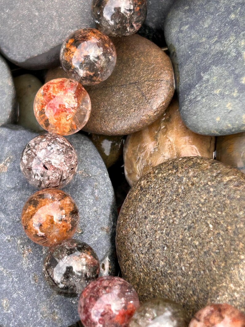 Puede incluir: Primer plano de varias piedras preciosas esf&eacute;ricas transparentes con inclusiones internas de color naranja, marr&oacute;n y negro. Las piedras preciosas est&aacute;n dispuestas entre cantos rodados lisos, grises y marrones, creando una est&eacute;tica natural y terrosa.