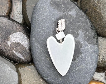 May include: A light green heart-shaped gua sha tool with a silver metal and crystal accent. The tool is resting on a dark gray stone, surrounded by other wet, smooth stones. The gua sha is a self-care beauty product.