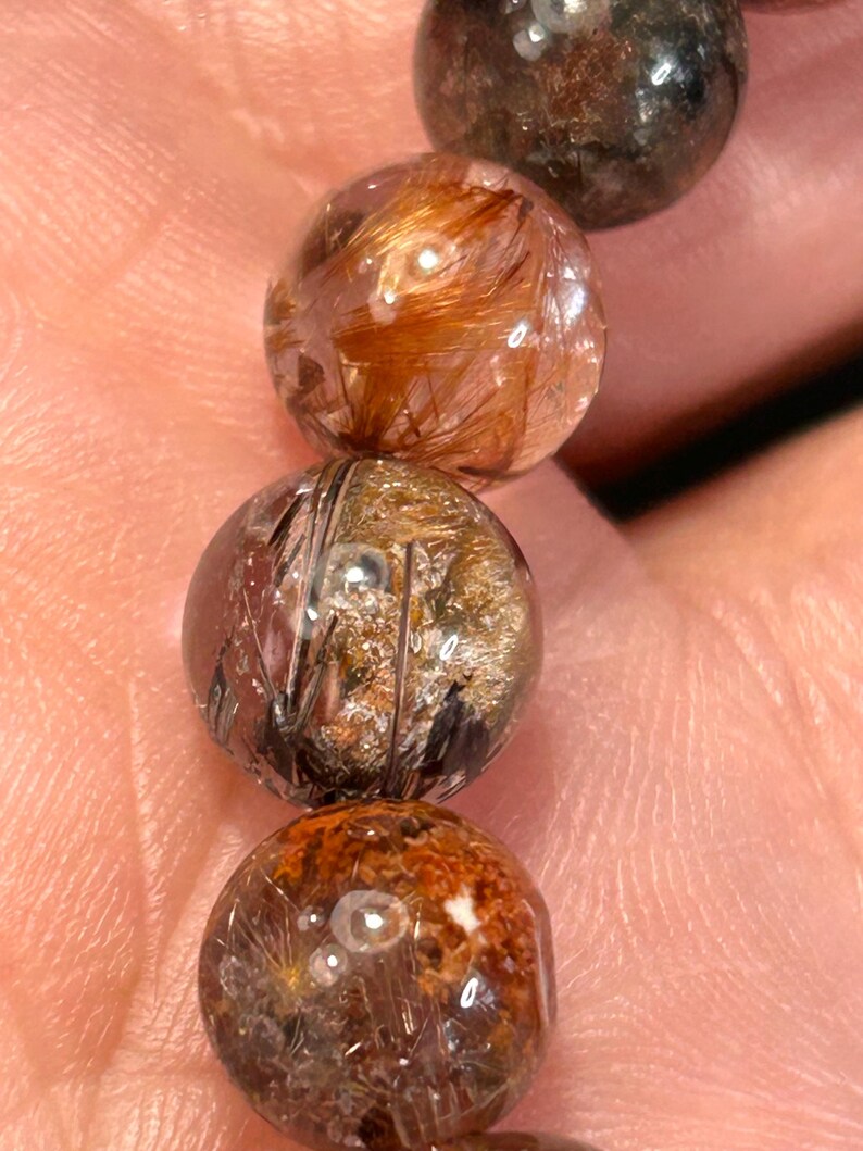 Puede incluir: Primer plano de una pulsera con varias cuentas redondas y transparentes. Cada cuenta tiene inclusiones marrones, doradas y negras, creando una apariencia texturizada &uacute;nica. Las cuentas est&aacute;n ensartadas, mostrando las variaciones naturales dentro de las piedras.