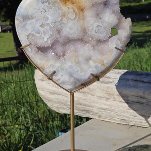 May include: A heart-shaped geode displayed on a gold-coloured metal stand. The geode has a mix of white, lavender, and golden-brown hues. The stand features a circular base with a floral design and a vertical support.