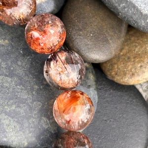 Puede incluir: Primer plano de un collar de cuentas redondas y transparentes con inclusiones de color marr&oacute;n rojizo. Las cuentas descansan sobre un lecho de piedras de r&iacute;o lisas, grises y marrones. Las cuentas tienen un aspecto pulido y vidrioso.
