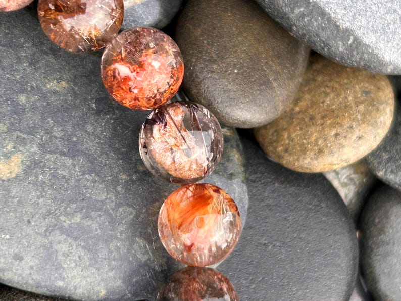 Puede incluir: Primer plano de un collar de cuentas redondas y transparentes con inclusiones de color marr&oacute;n rojizo. Las cuentas descansan sobre un lecho de piedras de r&iacute;o lisas, grises y marrones. Las cuentas tienen un aspecto pulido y vidrioso.