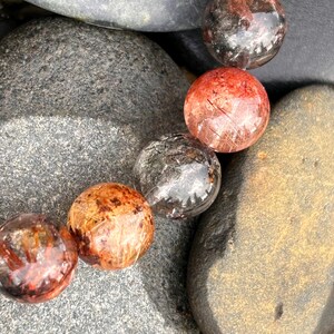 Puede incluir: Primer plano de una pulsera con varias cuentas redondas y transl&uacute;cidas con inclusiones de color marr&oacute;n rojizo y negro. Las cuentas descansan sobre un lecho de piedras lisas, grises y marrones, creando una est&eacute;tica natural y terrosa.