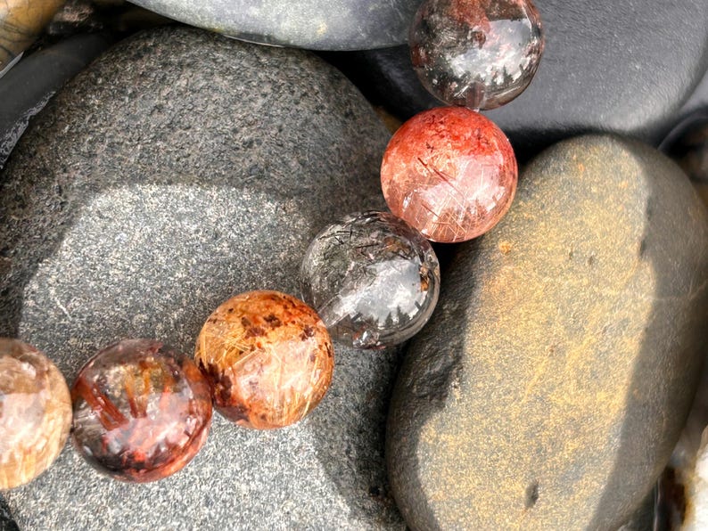 Puede incluir: Primer plano de una pulsera con varias cuentas redondas y transl&uacute;cidas con inclusiones de color marr&oacute;n rojizo y negro. Las cuentas descansan sobre un lecho de piedras lisas, grises y marrones, creando una est&eacute;tica natural y terrosa.