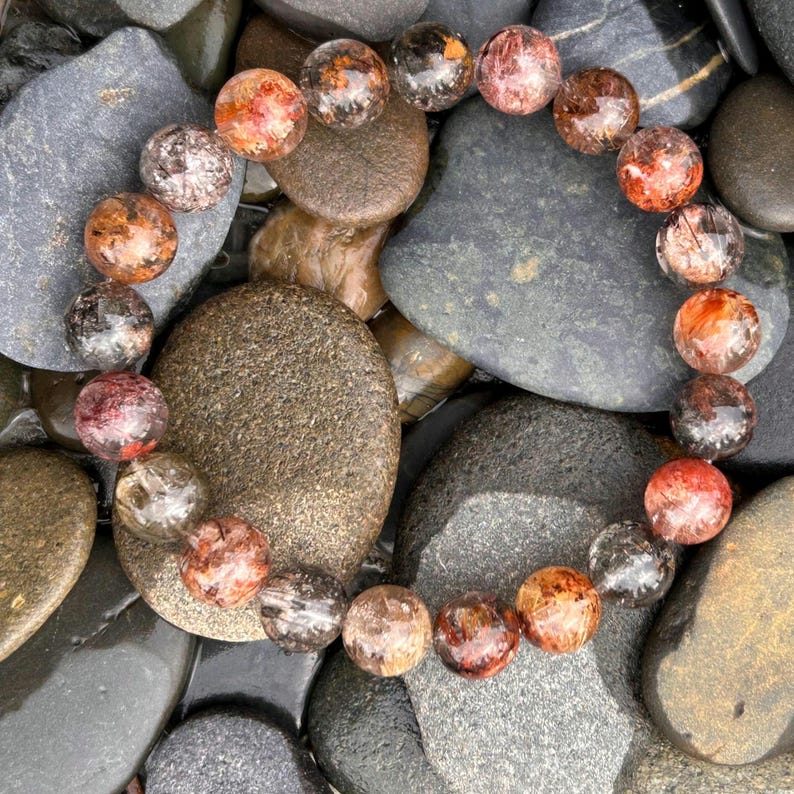 Puede incluir: Un brazalete de cuentas redondas y transl&uacute;cidas con inclusiones rojas, naranjas y negras. Las cuentas se exhiben sobre un lecho de piedras de r&iacute;o lisas, grises y marrones, creando una est&eacute;tica natural.