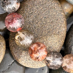 Puede incluir: Primer plano de un collar de cuentas redondas y transparentes con inclusiones rojas, marrones y negras. Las cuentas descansan sobre una colecci&oacute;n de piedras lisas grises y marrones. Las cuentas tienen un di&aacute;metro de aproximadamente 2 cm.