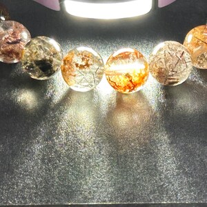 Puede incluir: Primer plano de una pulsera con cuentas redondas y transparentes con inclusiones internas de minerales rojizos y negros. Las cuentas est&aacute;n iluminadas, creando reflejos y sombras sobre una superficie oscura. La pulsera es una joya.