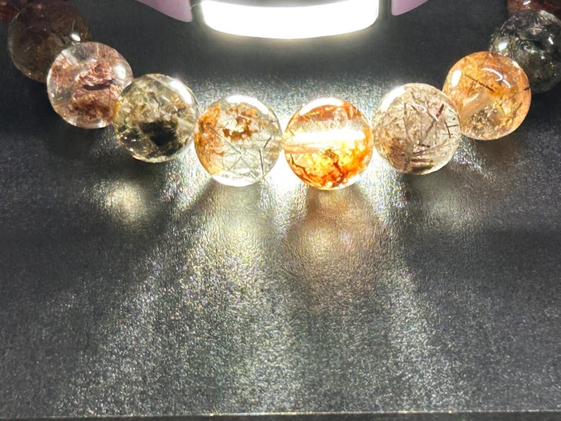 Puede incluir: Primer plano de una pulsera con cuentas redondas y transparentes con inclusiones internas de minerales rojizos y negros. Las cuentas est&aacute;n iluminadas, creando reflejos y sombras sobre una superficie oscura. La pulsera es una joya.