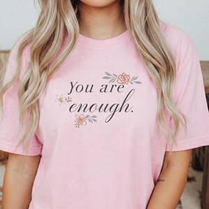 Può includere: T-shirt rosa chiaro con la scritta "You are enough" in corsivo, ornata da motivi floreali. La maglietta a maniche corte è di colore uniforme.