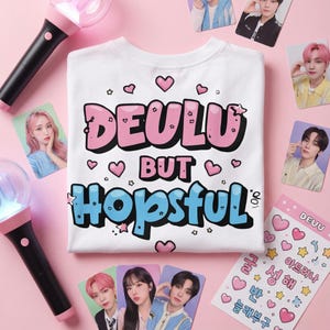 May include: A white t-shirt with the text "DEULU BUT Hopsful" in pink and blue, decorated with hearts and stars. The image also features photo cards, stickers, and two pink and black light sticks.