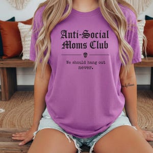May include: A purple t-shirt with the text "Anti-Social Moms Club" in a gothic font, with a skull graphic. Below the text is the phrase "We should hang out never."