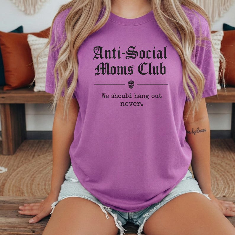 May include: A purple t-shirt with the text "Anti-Social Moms Club" in a gothic font, with a skull graphic. Below the text is the phrase "We should hang out never."