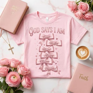 Può includere: Maglietta rosa chiaro con la scritta "God Says I Am" e nastri con le parole "Loved", "Worthy", "Blessed", "Strong", "Chosen" e "Beautiful". L'immagine include una Bibbia, una croce, rose e caffè.