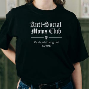 May include: Black t-shirt with the text "Anti-Social Moms Club" in a gothic font. Below the text is a skull graphic and the phrase "We should hang out never."