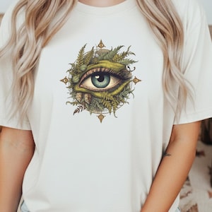 Può includere: T-shirt color crema con una grafica dettagliata di un occhio con iride blu, circondato da fogliame verde. Il disegno dell'occhio è centrato sulla maglietta e incorniciato da un disegno a forma di bussola con accenti dorati. La maglietta è in materiale morbido.