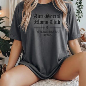 May include: Dark gray t-shirt with the text "Anti-Social Moms Club" in a gothic font. Below the text is a skull graphic and the phrase "We should hang out never."