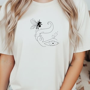 May include: Cream-colored t-shirt featuring a minimalist black line art design. The design includes a stylized eye, a bee, and flowing lines. The t-shirt is made of a soft, comfortable material.
