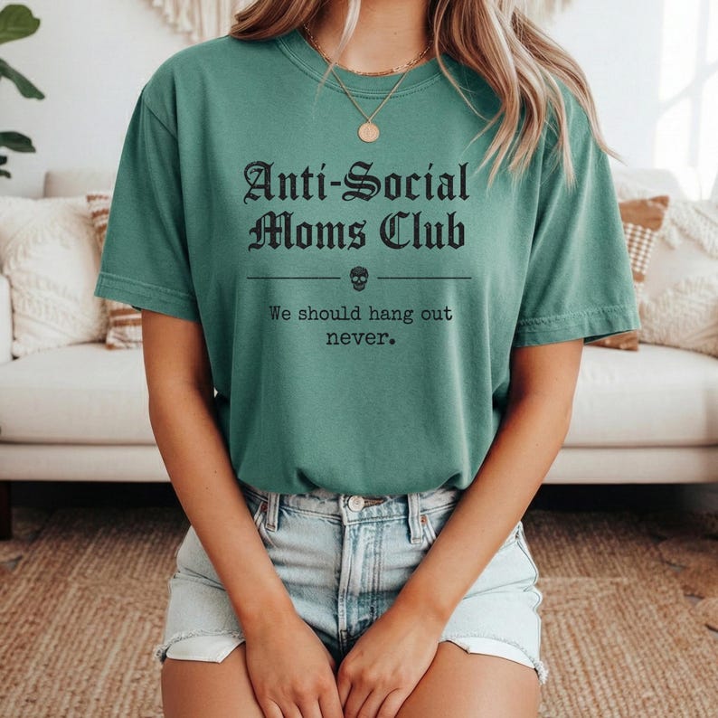 May include: A teal green t-shirt with the text "Anti-Social Moms Club" in a gothic font. Below the text is a skull graphic and the phrase "We should hang out never."