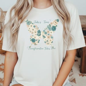 Può includere: T-shirt color crema con un motivo a farfalla floreale nei toni del blu, crema e bianco. Il testo "Takes Time" e "Transformation Takes Time" è stampato sopra e sotto la grafica della farfalla.