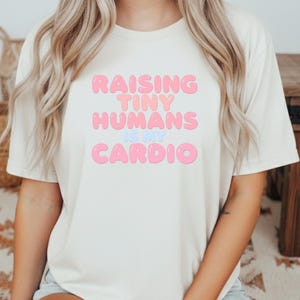 Può includere: T-shirt color crema con la scritta "RAISING TINY HUMANS IS MY CARDIO" in lettere rosa e azzurre. La maglietta ha maniche corte e girocollo.