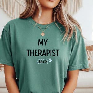 Puede incluir: Camiseta de color verde azulado con el texto "MY THERAPIST SAID" en letras negras. La palabra "SAID" está dentro de una nube blanca. La camiseta es de manga corta y cuello redondo.