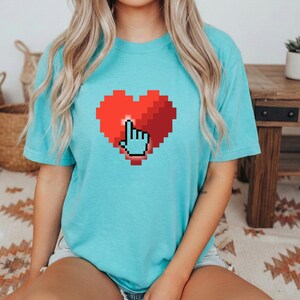 Può includere: T-shirt azzurra con un cuore rosso pixelato e un cursore a mano pixelato. La grafica è centrata sulla parte anteriore della maglietta. La maglietta ha maniche corte e girocollo.