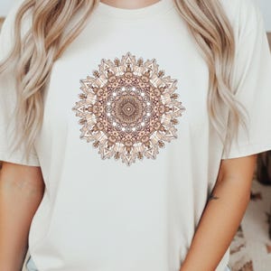 Può includere: T-shirt color crema con un design mandala dettagliato nei toni del marrone e beige. L'intricato motivo circolare è centrato sulla maglietta, creando un design simmetrico. La maglietta ha maniche corte e girocollo.