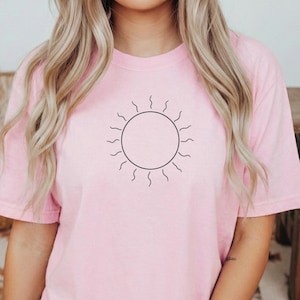 Può includere: Una maglietta rosa chiaro con un disegno di sole nero. La grafica del sole è un semplice contorno con un centro circolare e linee ondulate che si estendono verso l'esterno. La maglietta è realizzata in materiale morbido.