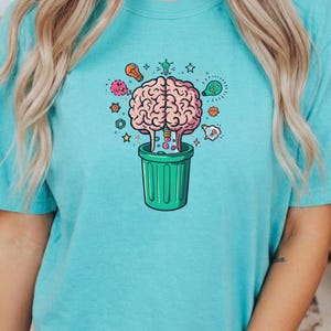 Può includere: T-shirt azzurra con una grafica colorata di un cervello rosa che esce da un cestino verde. Il design include stelle, lampadine e altri elementi stravaganti. La maglietta è realizzata in materiale morbido.