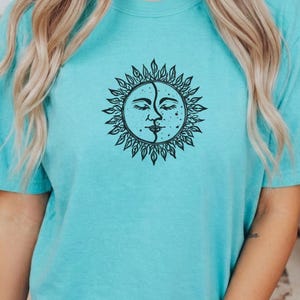 Può includere: Una maglietta azzurra con una grafica nera di sole e luna. Il design mostra un volto di sole e luna con gli occhi chiusi e un viso a pois. Il sole e la luna sono circondati da un disegno a forma di foglia.