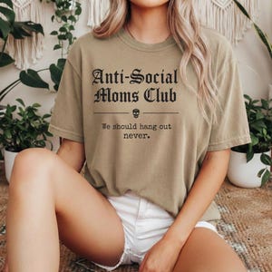 May include: Olive green t-shirt with the text "Anti-Social Moms Club" in a gothic font, a skull graphic, and the phrase "We should hang out never." The shirt is a casual, comfortable style.