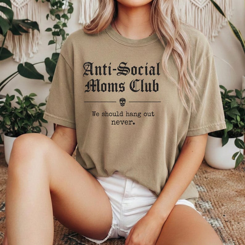 May include: Olive green t-shirt with the text "Anti-Social Moms Club" in a gothic font, a skull graphic, and the phrase "We should hang out never." The shirt is a casual, comfortable style.