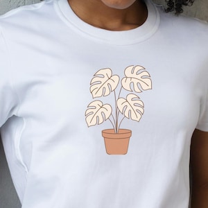 Può includere: T-shirt bianco con un'illustrazione minimalista di una pianta in vaso. La pianta ha grandi foglie stilizzate di colore beige chiaro e il vaso è marrone caldo. Il disegno è centrato sulla maglietta.