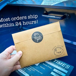May include: A brown envelope with a black "thank you" sticker and a "Happy Turtle Greetings" stamp is placed in a blue mailbox. The text "Most orders ship within 24 hours!" is visible.
