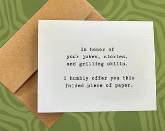 May include: A white folded card with black text, resting on a brown envelope. The card reads, "In honor of your jokes, stories, and grilling skills, I humbly offer you this folded piece of paper."