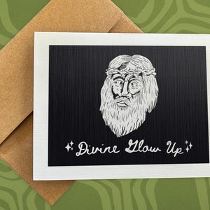 May include: Greeting card with a black background and a white illustration of a bearded figure wearing a crown of thorns. The text "Divine Glow Up" is written in white script below the illustration. The card is on a brown envelope.