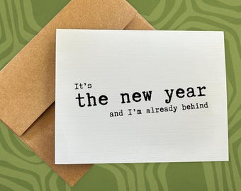 May include: A white greeting card with the text "It's the new year and I'm already behind" in black font. The card is paired with a brown envelope, set against a green patterned background.