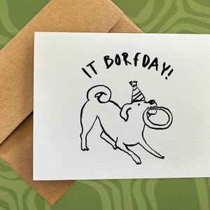 May include: A white greeting card with a black line drawing of a dog wearing a party hat and holding a ring in its mouth. The text "IT BORFDAY!" is above the dog. The card is paired with a brown envelope.