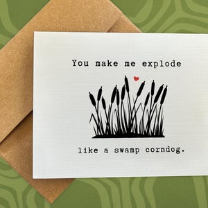 May include: A white greeting card with the text "You make me explode like a swamp corndog." A red heart is above a black illustration of cattails. The card is inside a brown envelope.