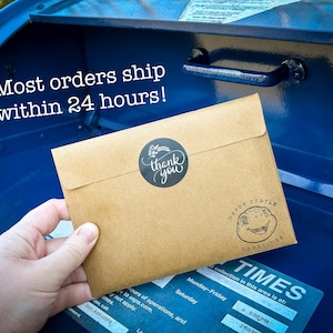 May include: A brown paper envelope with a "thank you" sticker and a "Happy Turtle Greetings" stamp. The envelope is being placed in a blue mailbox. The text "Most orders ship within 24 hours!" is visible on the mailbox.