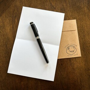 May include: A blank white card with a black pen on top of it. The card is folded in half. A brown envelope with the text "Happy Turtle Greetings" is in the background.