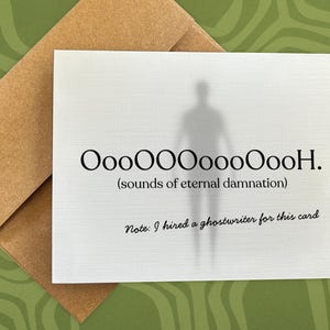 May include: A white greeting card with a brown envelope featuring a faint gray silhouette of a figure. The text reads OooOOOOoooOooH. (sounds of eternal damnation) and Note: I hired a ghostwriter for this card in black serif and script fonts.