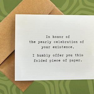May include: A white greeting card with black text, resting on a brown envelope. The card reads, "In honor of the yearly celebration of your existence, I humbly offer you this folded piece of paper." The background is a green, patterned surface.