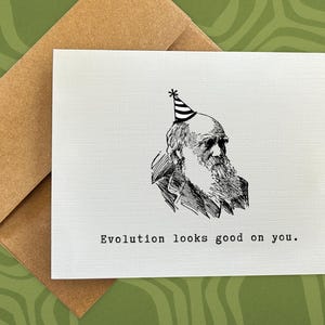 May include: A white card with a black and white illustration of Charles Darwin wearing a party hat. The text on the card reads "Evolution looks good on you."