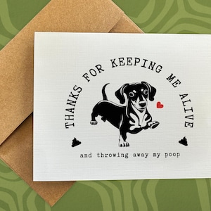 May include: A white card with black text that reads "Thanks for keeping me alive and throwing away my poop." A black silhouette of a dachshund is in the center of the card with a red heart above it.