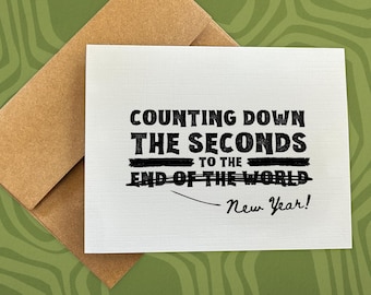 May include: A white greeting card with the text "COUNTING DOWN THE SECONDS TO THE END OF THE WORLD" in black, with "New Year!" written below. The card is inside a brown envelope, set against a green patterned background.